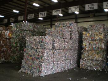 Bundles of recycled material stacked in a warehouse under signs indicating what they are.